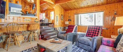 A cozy cabin interior with wooden walls, a dining area, and a living room with a couch and chairs.