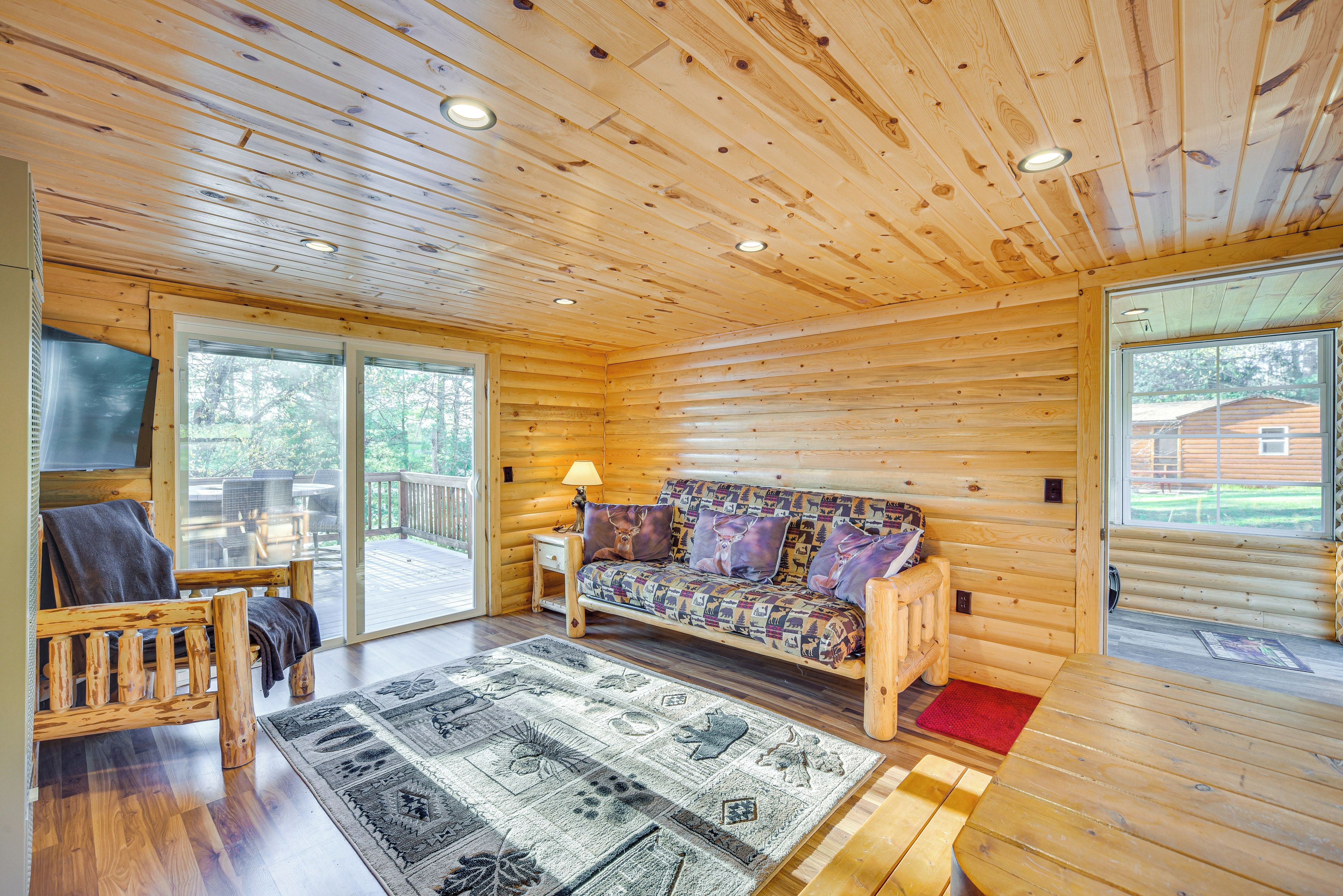 A log cabin interior with a wooden ceiling, a sofa with patterned cushions, a wooden bench, a rug with animal prints, and a sliding glass door leading to an outdoor deck.