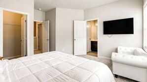A bedroom with a large bed, a white armchair, a flat-screen TV mounted on the wall, and multiple doors leading to other rooms. - Lawrenceville Townhome w/ Community Pool Access! (Lawrenceville)