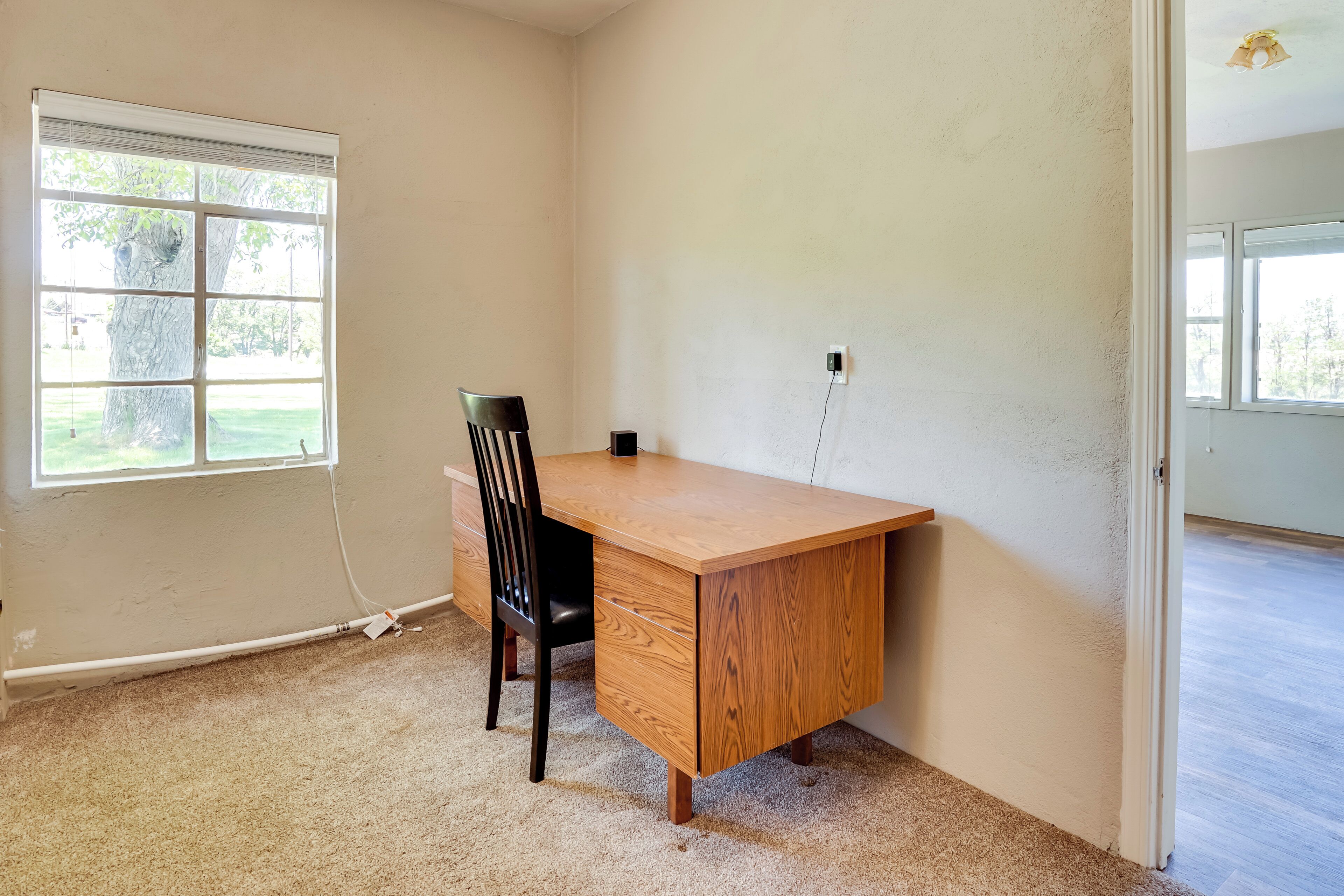 A room with a wooden desk, a black chair, a window with blinds, and a carpeted floor.
