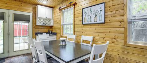 Interior de una cabaña de madera con una mesa de comedor, sillas blancas, una zona de cocina y un cuadro en la pared.