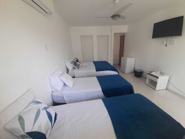 Une chambre d’hôtel avec trois lits simples, un ventilateur de plafond, une télévision et un petit bureau avec un réfrigérateur.