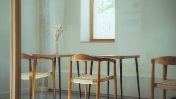 A dining area with wooden chairs and a table, a vase with dried flowers, and a window with a view of greenery.