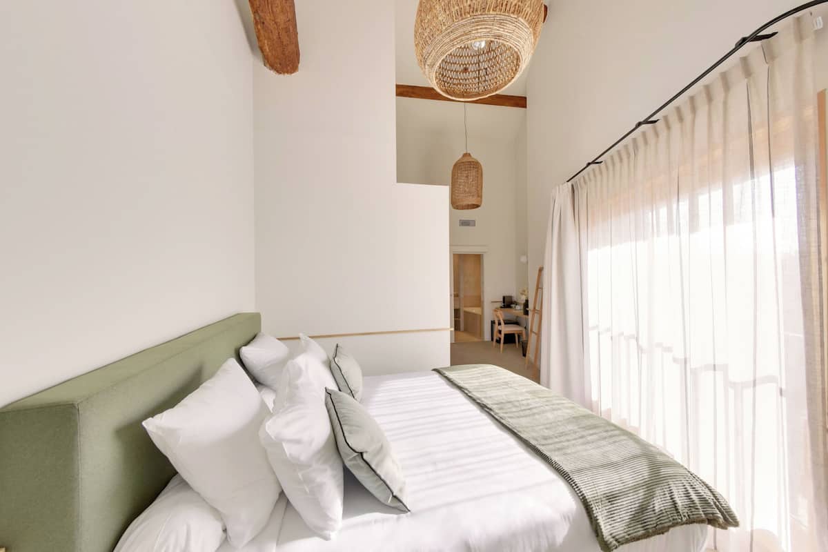 Un dormitorio con cabecera verde, ropa de cama blanca, una lámpara de fibra natural y vistas a un baño a través de una ventana con cortinas translúcidas.