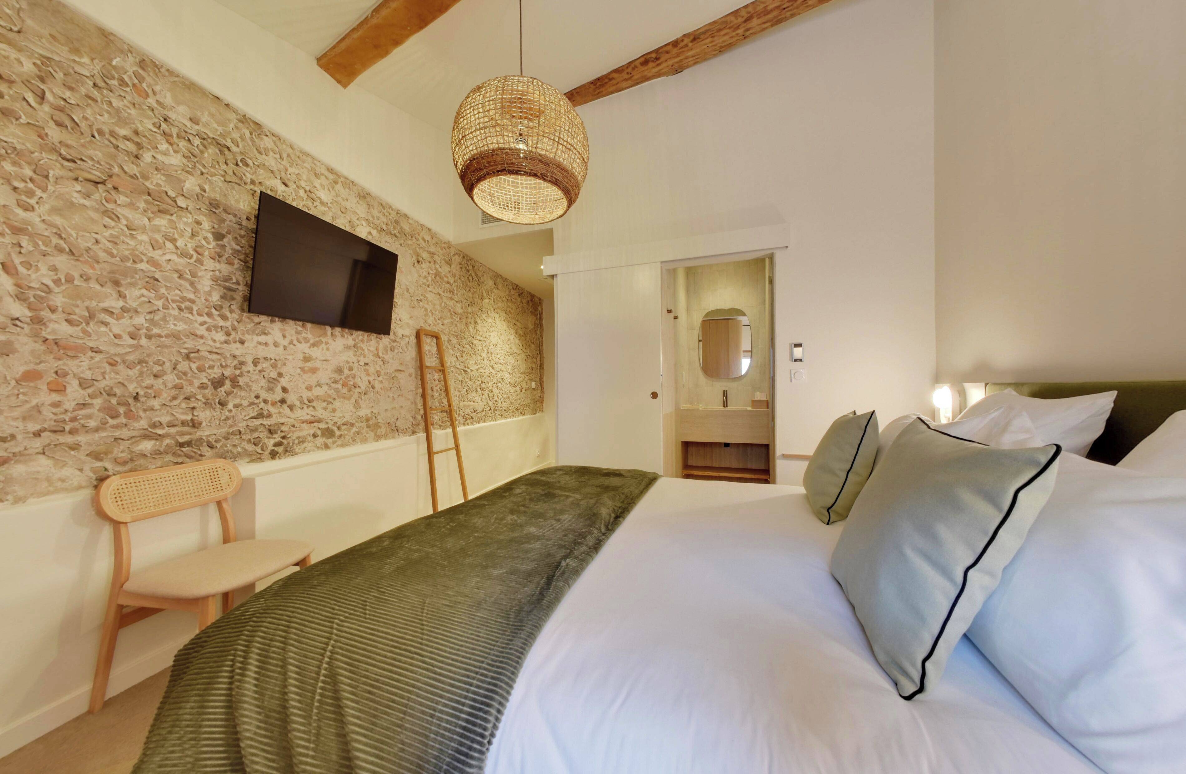 A bedroom with a stone wall, a bed with white and green bedding, a wooden chair, a woven light fixture, and a flat-screen TV mounted on the wall.