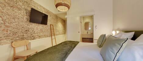 A bedroom with a stone wall, a bed with white and green bedding, a wooden chair, a woven light fixture, and a flat-screen TV mounted on the wall.