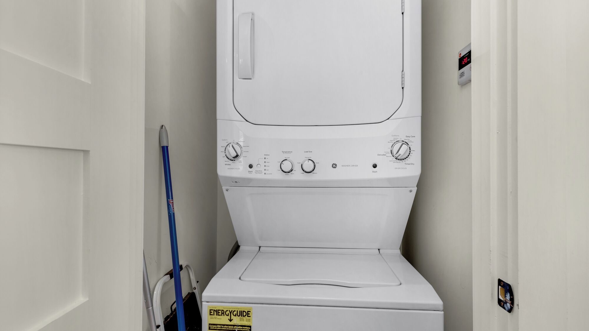 A front-loading washing machine in a laundry room with a door, a mop, and an ENERGY GUIDE sticker.