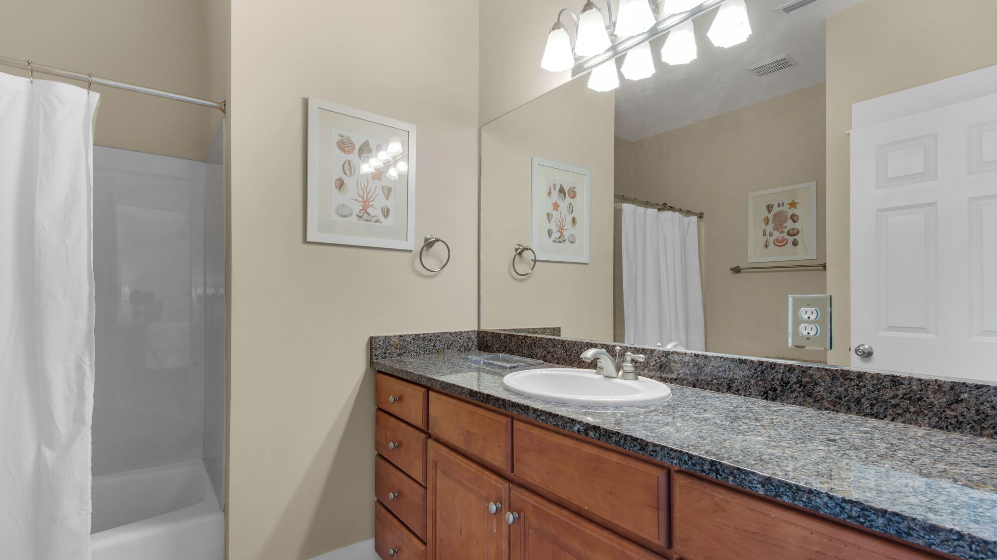 A bathroom with a granite countertop, a sink, wooden cabinets, a large mirror, and framed artwork on the wall.