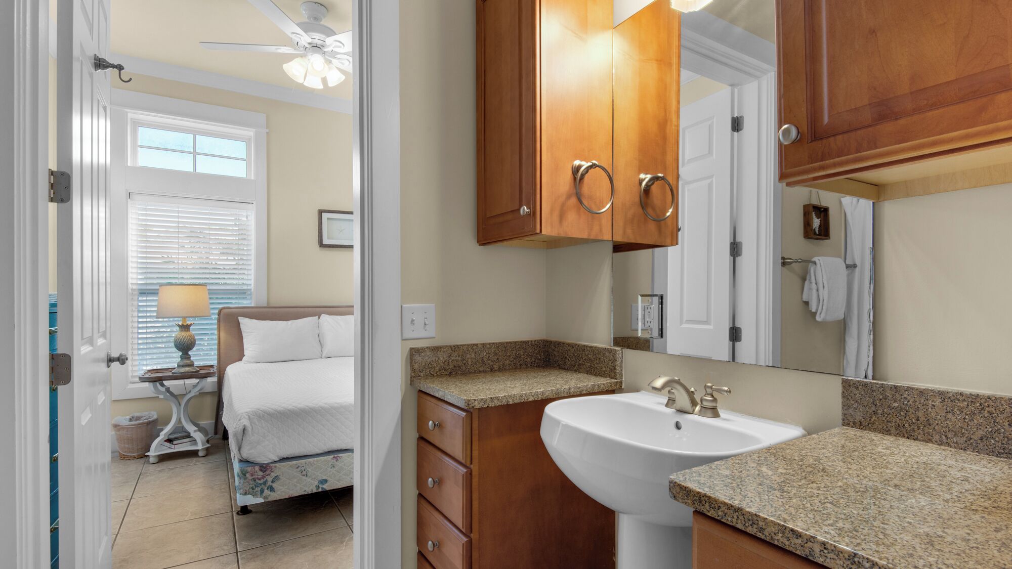 A hotel bathroom with a sink, mirror, wooden cabinets, and a bed in the adjacent room.