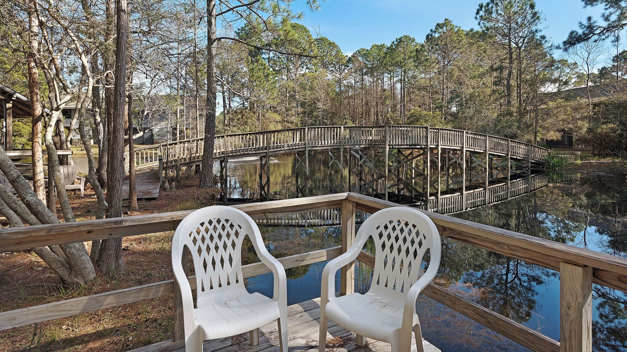 A wooden deck with two white plastic chairs overlooking a calm body of water, surrounded by trees and a wooden bridge.