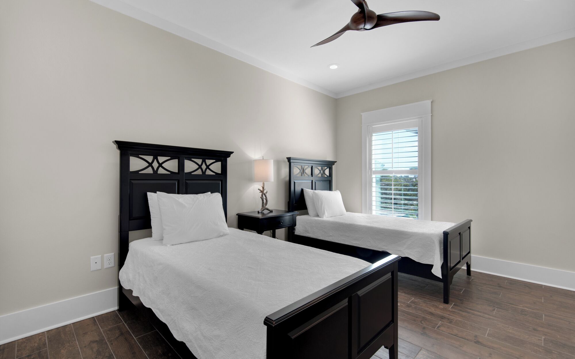 A hotel room with two beds, a ceiling fan, a window with blinds, and a nightstand with a lamp.