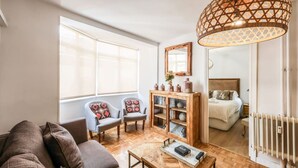 Een gezellige woonkamer met een bank, fauteuils, een bijzettafeltje en een houten kast. Een groot raam zorgt voor natuurlijk licht en uitzicht op de slaapkamer.