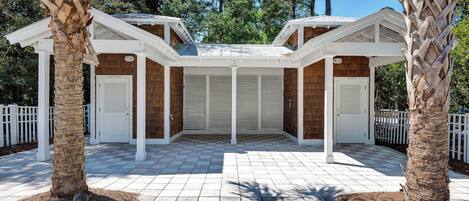 A covered outdoor area with two doors labeled 'Gentlemen' and 'Ladies,' surrounded by palm trees and a white fence.
