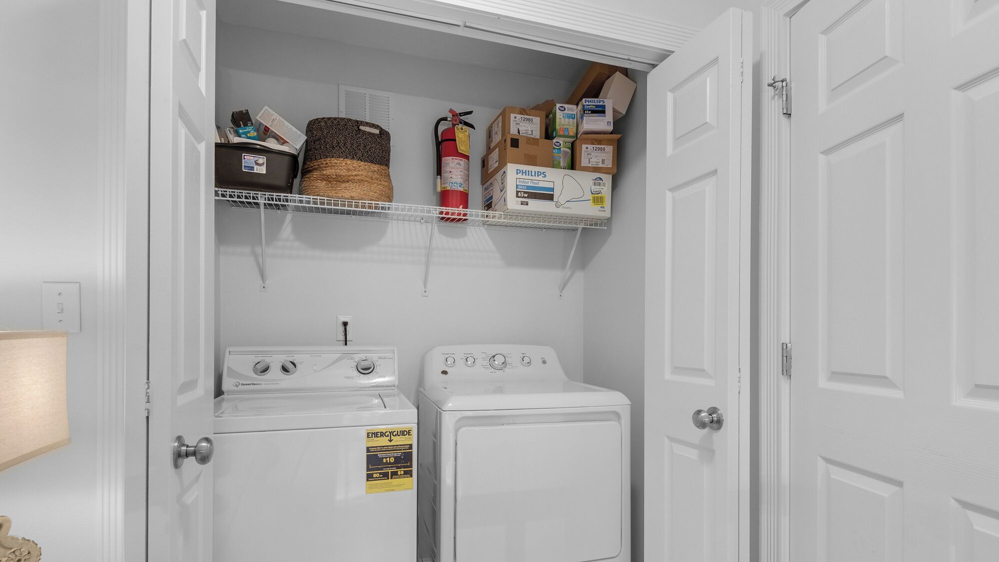 A laundry room with two stacked washers and dryers, a fire extinguisher, and shelves above holding boxes and a basket.