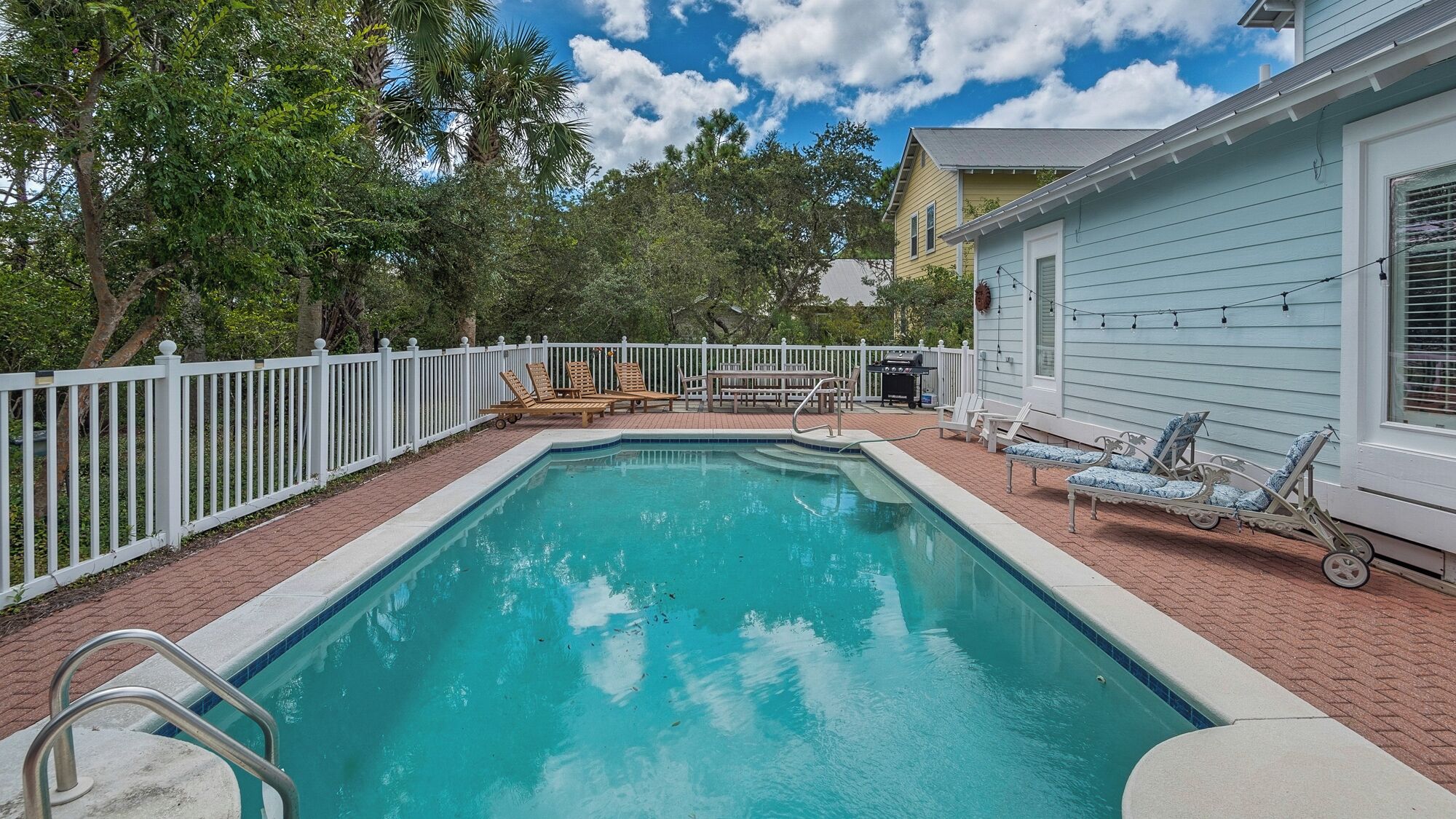 A rectangular swimming pool with a white deck and a white fence surrounding it. There are lounge chairs and a barbecue grill on the deck.