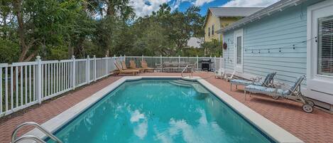 A rectangular swimming pool with a white deck and a white fence surrounding it. There are lounge chairs and a barbecue grill on the deck.