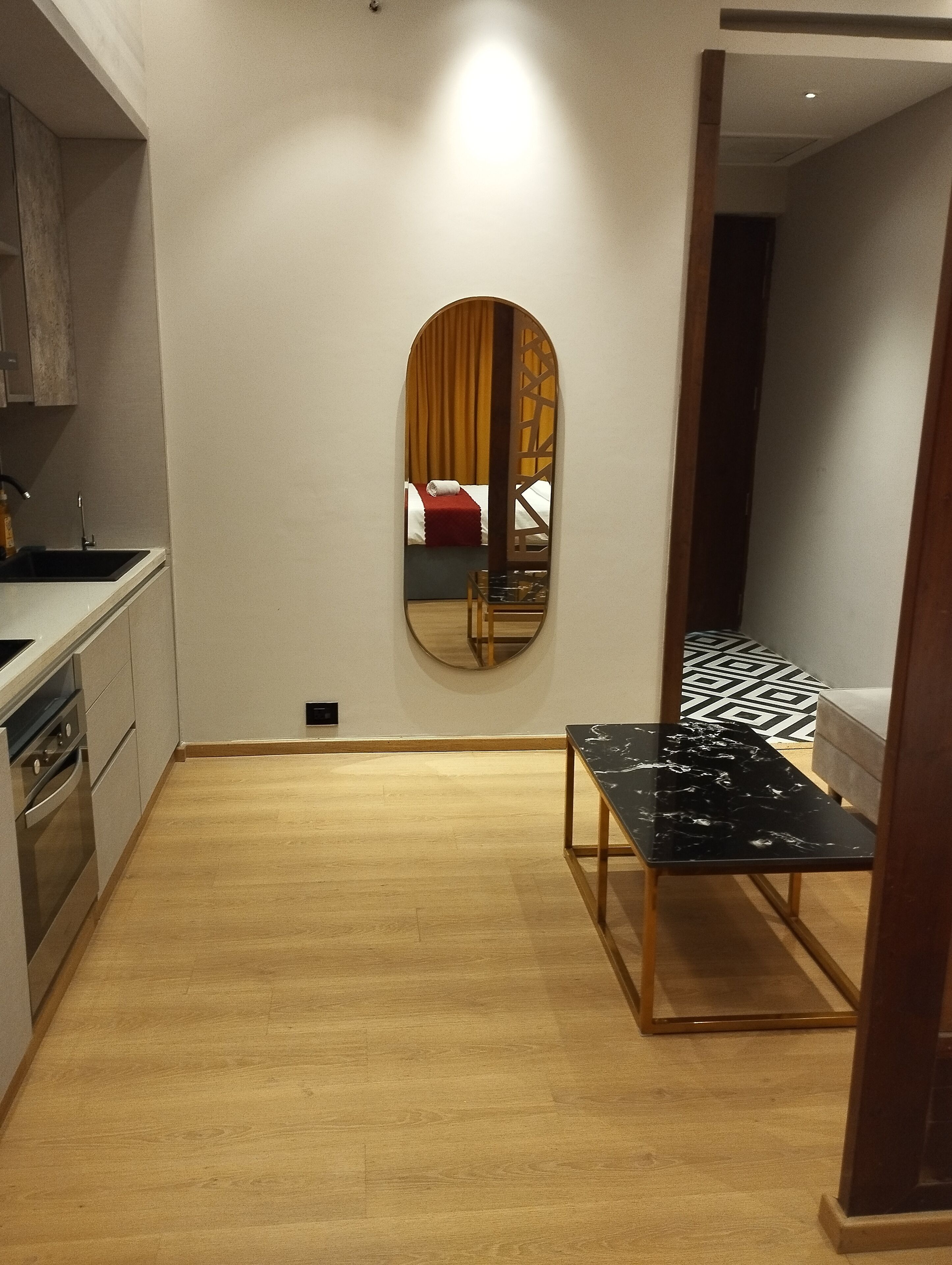 A compact kitchen with modern appliances, a dining area with a black marble table, and a mirrored wall reflecting a bedroom with a bed and curtains.