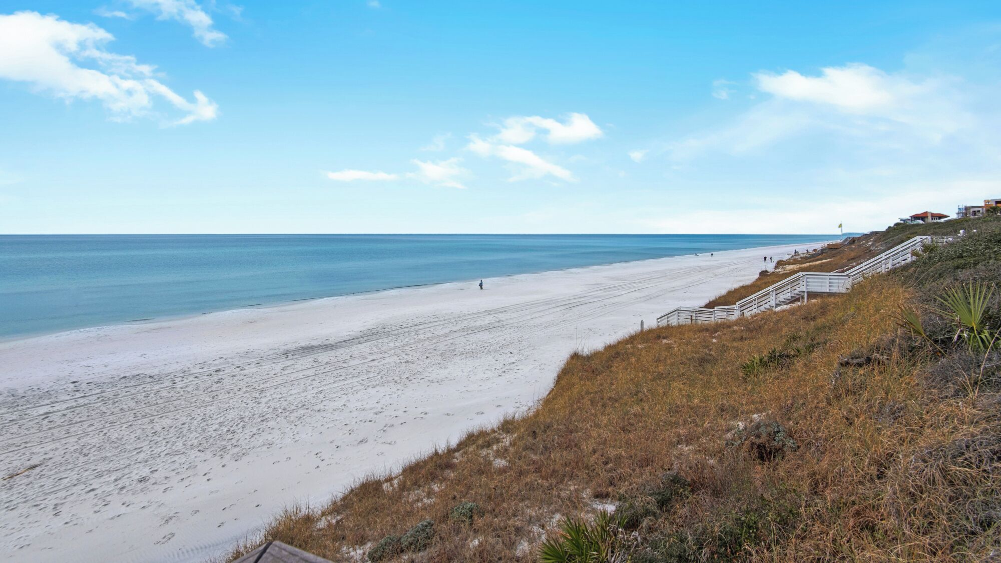 Une plage avec un escalier descendant vers le rivage, un ciel dégagé et quelques personnes visibles au loin.
