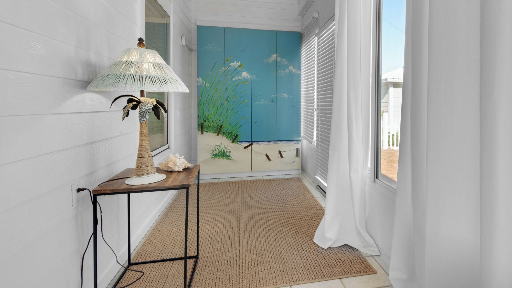 Um corredor com luminária, uma mesa pequena, um painel decorativo com cena de praia e vista pela janela.