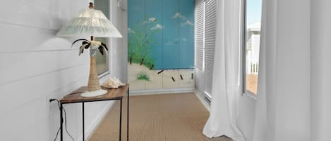 Um corredor com luminária, uma mesa pequena, um painel decorativo com cena de praia e vista pela janela.