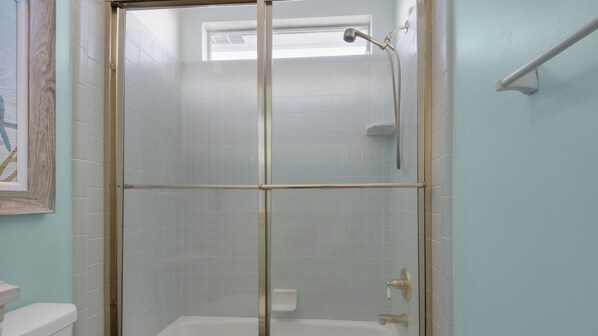 Cabina doccia con pareti in vetro, soffione a mano, water, vaso igienico bianco e una piccola finestra sopra la doccia.
