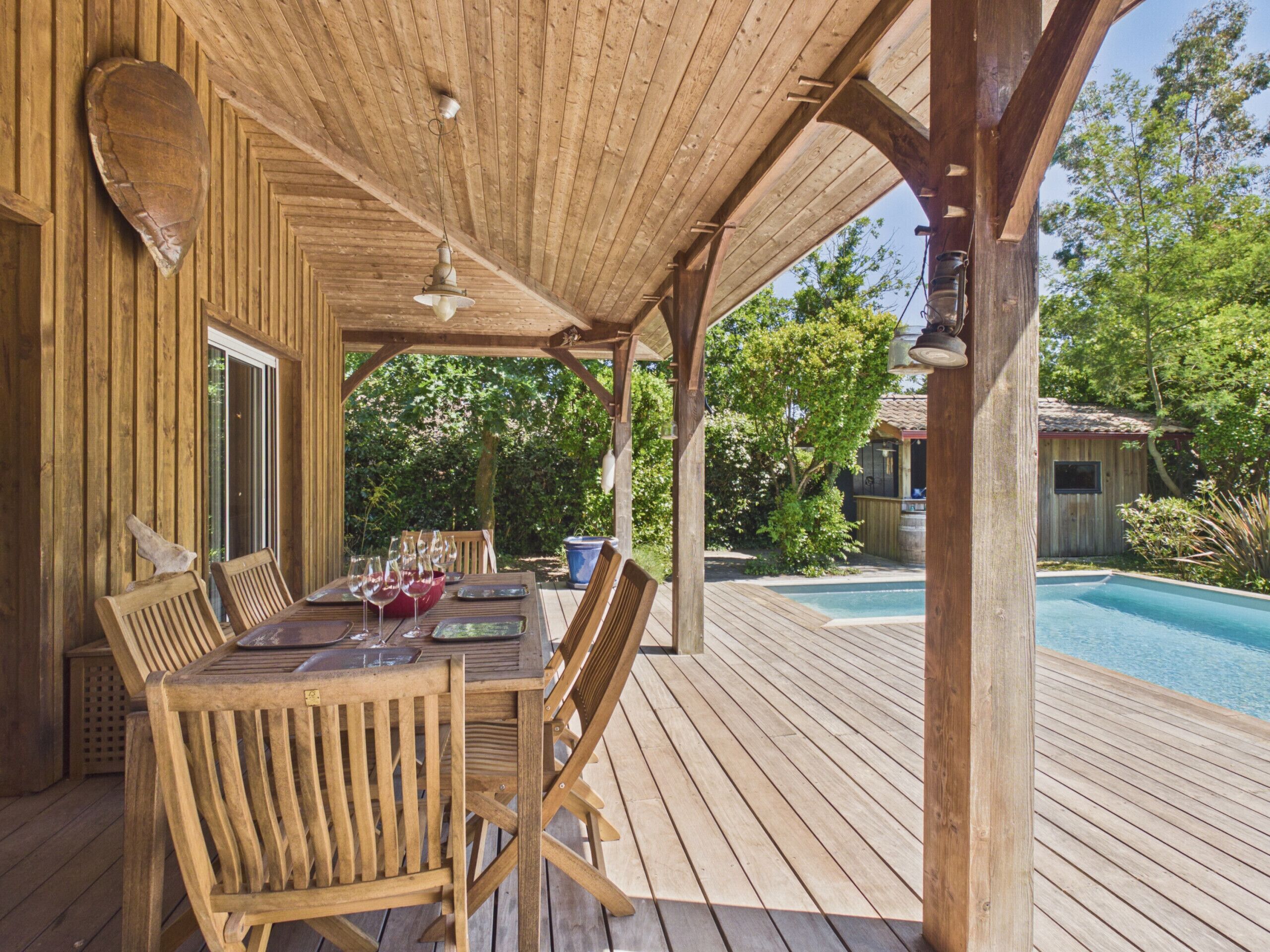 Une terrasse en bois avec une table dressée pour le repas, entourée de chaises, et une piscine en arrière-plan.