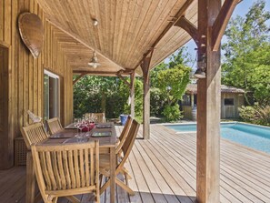 Una terraza de madera con una mesa lista para comer, rodeada de sillas, y una piscina al fondo.