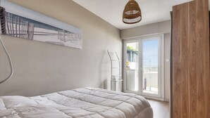 A bedroom with a large bed, a wooden wardrobe, a white radiator, and a view of the outside through a sliding glass door. - Flat for 4, sea view and right on the water (Saint-Hilaire-de-Riez)