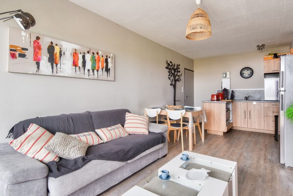 Ein modernes Wohnzimmer mit grauem Sofa, einem Couchtisch und einem Essbereich mit Tisch und Stühlen. Im Hintergrund ist eine Küche mit Holdekor und Geräten zu sehen.