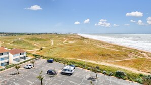 A parking lot with several cars and a caravan, a building with a red roof, and a beach with a sandy path leading to the water. - Flat for 4, sea view and right on the water (Saint-Hilaire-de-Riez)