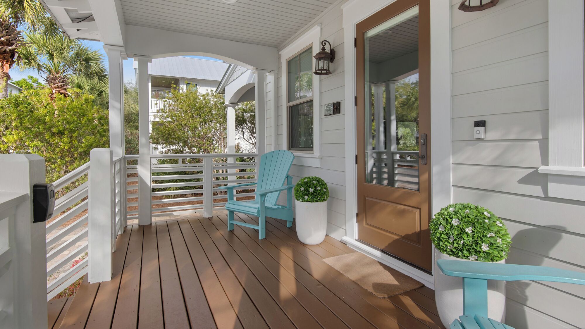 A covered porch with wooden flooring, a blue Adirondack chair, a potted plant, and a door with a window.