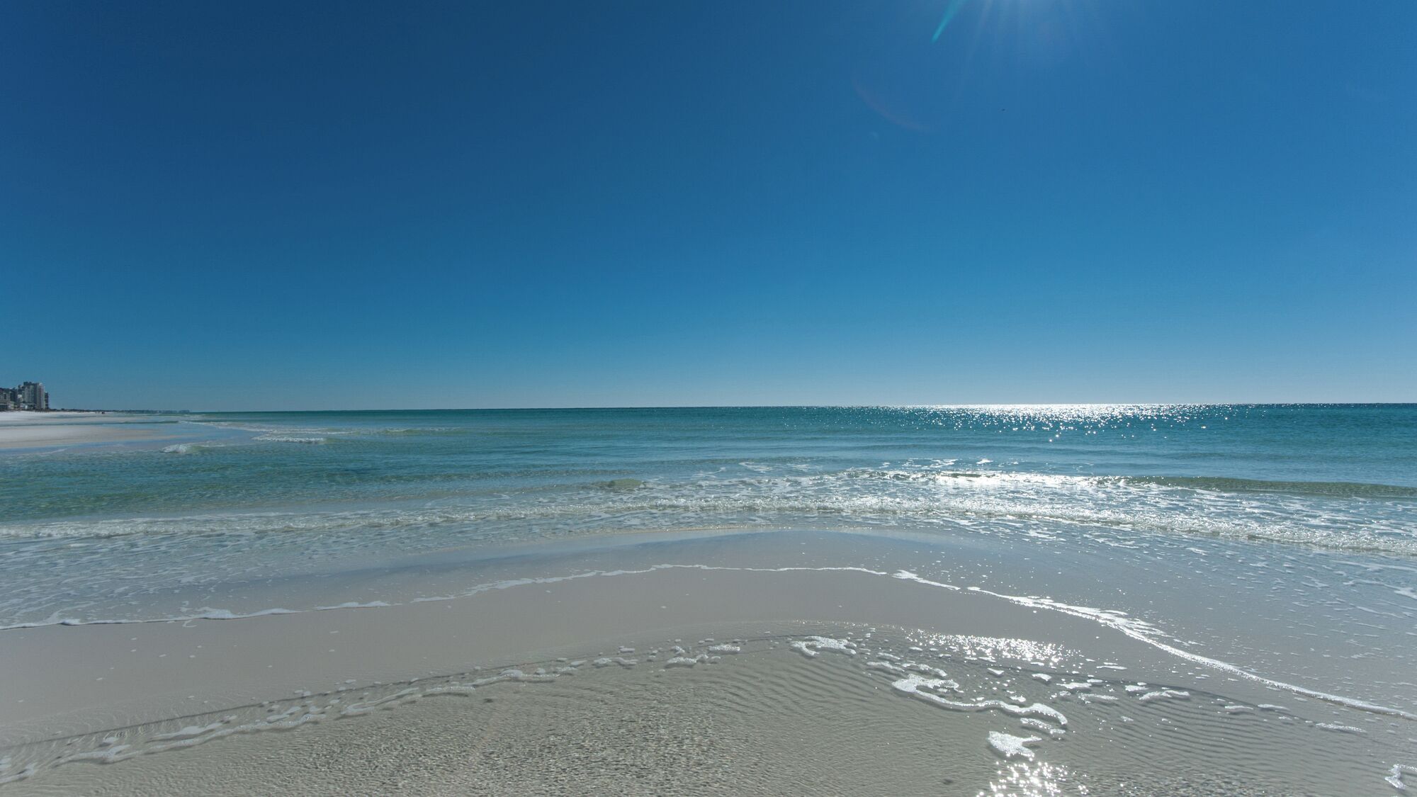 A beach with clear blue water, a sandy shore, and a distant building.
