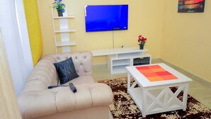 A living room with a beige sofa, a white coffee table, a flat-screen TV mounted on the wall, and a small shelf with a plant. - Lux Suites Rubi Homes Kisii (Kisii)