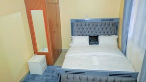 A bedroom with a bed, a wooden wardrobe, a small white cabinet, and a blue rug. - Lux Suites Rubi Homes Kisii (Kisii)