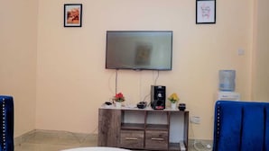 A living room with a flat-screen TV mounted on the wall, a wooden entertainment unit with electronic devices, and two blue chairs. - Lux Suites Rubi Homes Kisii (Kisii)