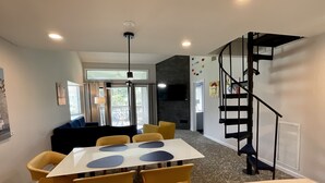 Zona pranzo moderna con tavolo bianco e sedie gialle, una scala nera e vista sull'esterno tramite porte in vetro.