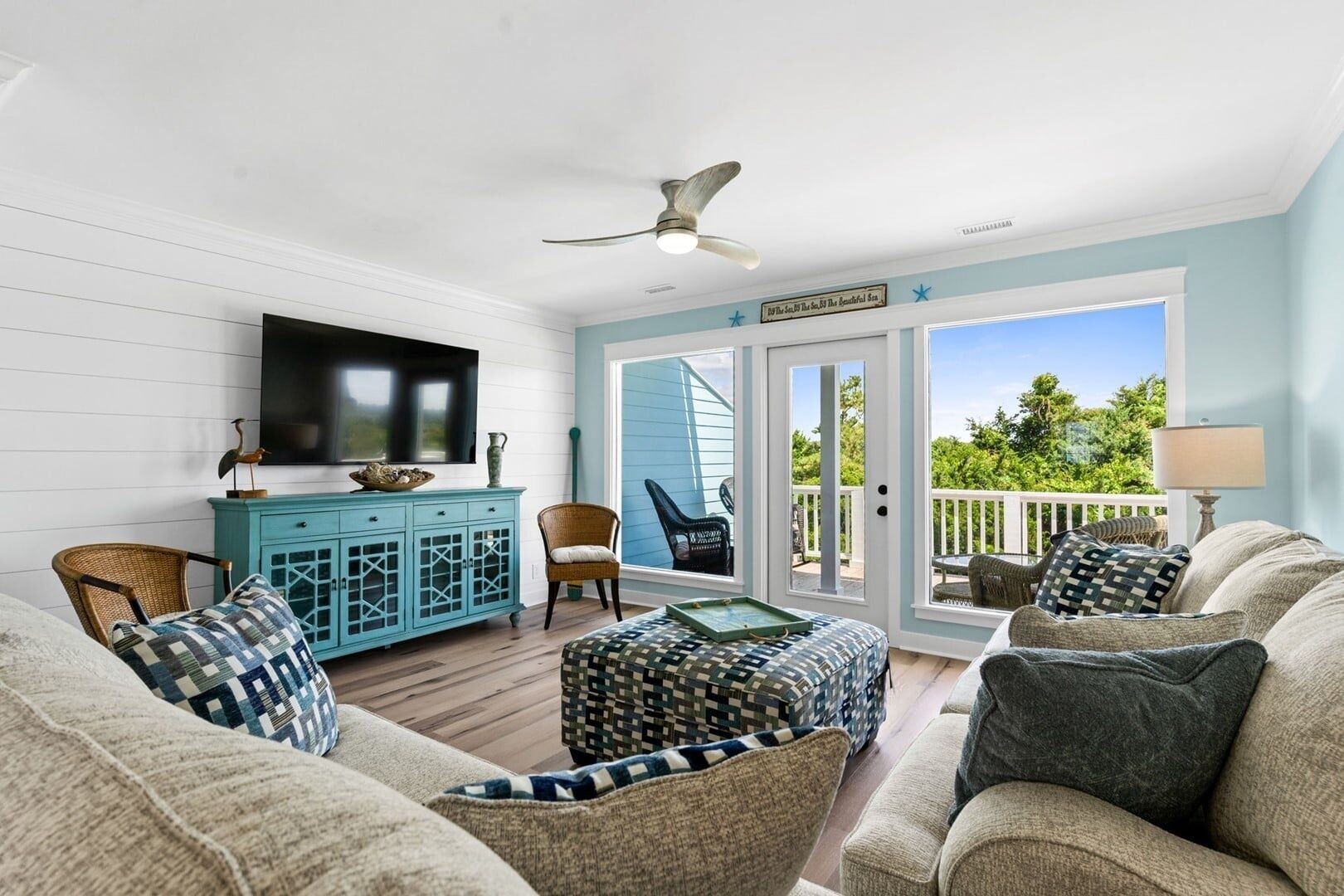 A spacious living area with a ceiling fan, a television, a blue cabinet, wicker furniture, and a view through the glass doors.