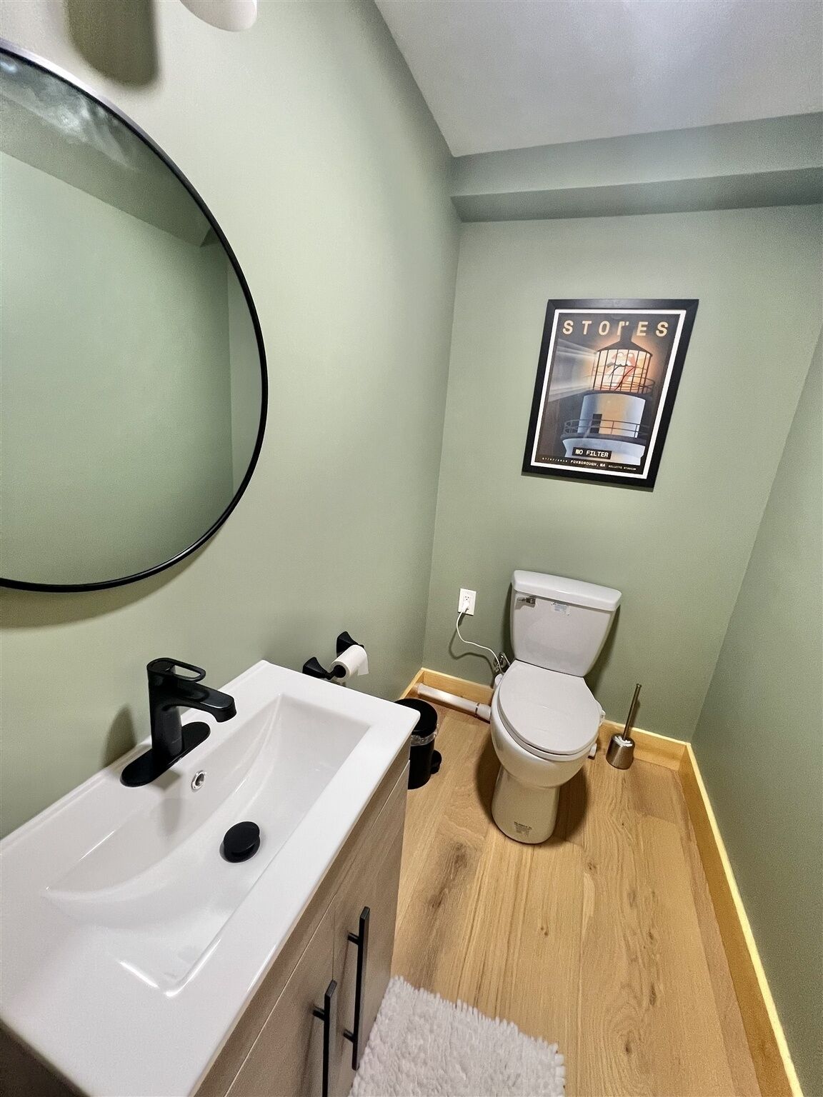 A bathroom with a white sink, a toilet, and a mirror. There is a framed picture on the wall and a white rug on the floor.
