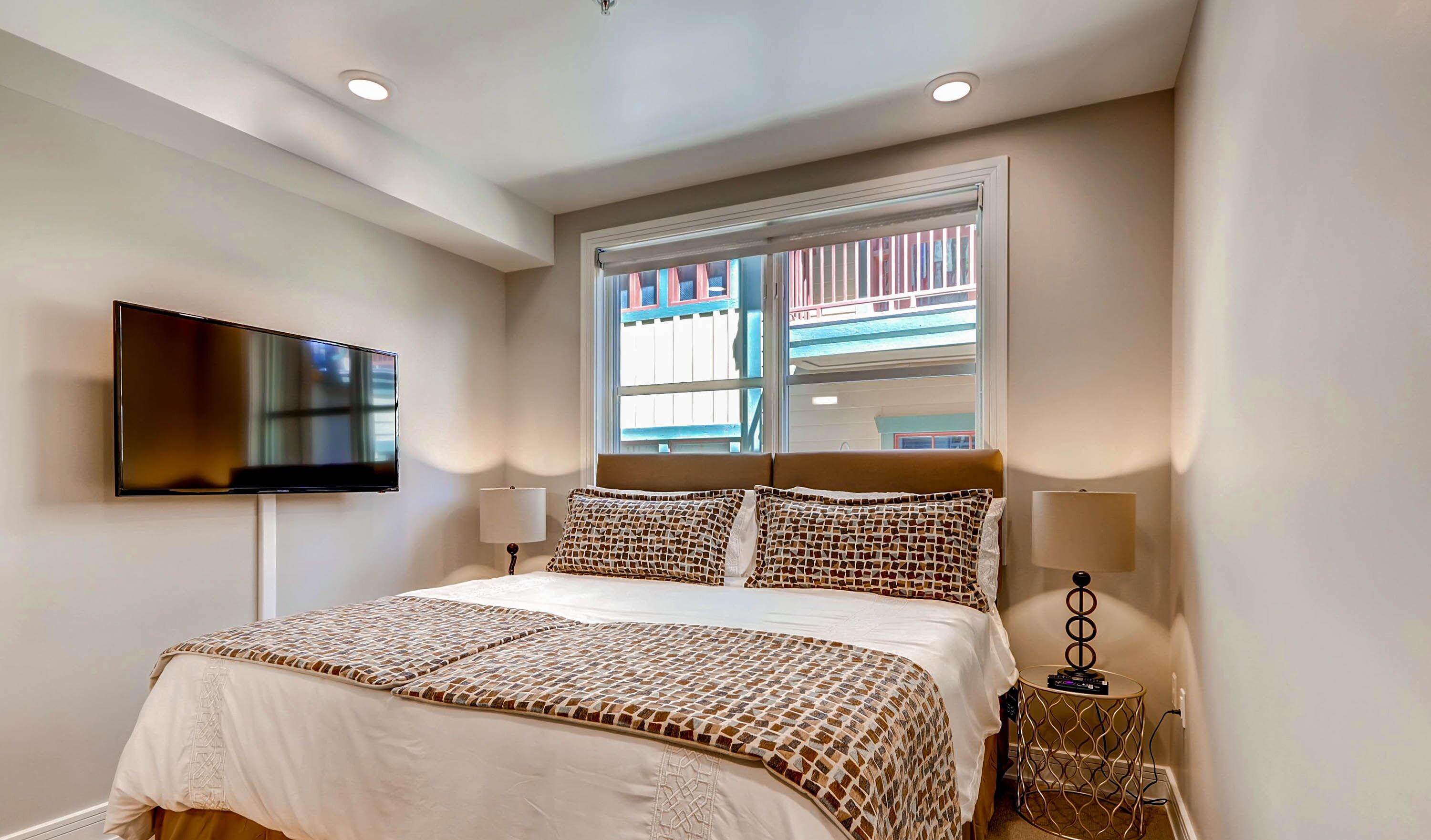 Un dormitorio con una cama, un televisor montado en la pared, una ventana con vistas a edificios y una mesita de noche con lámpara.