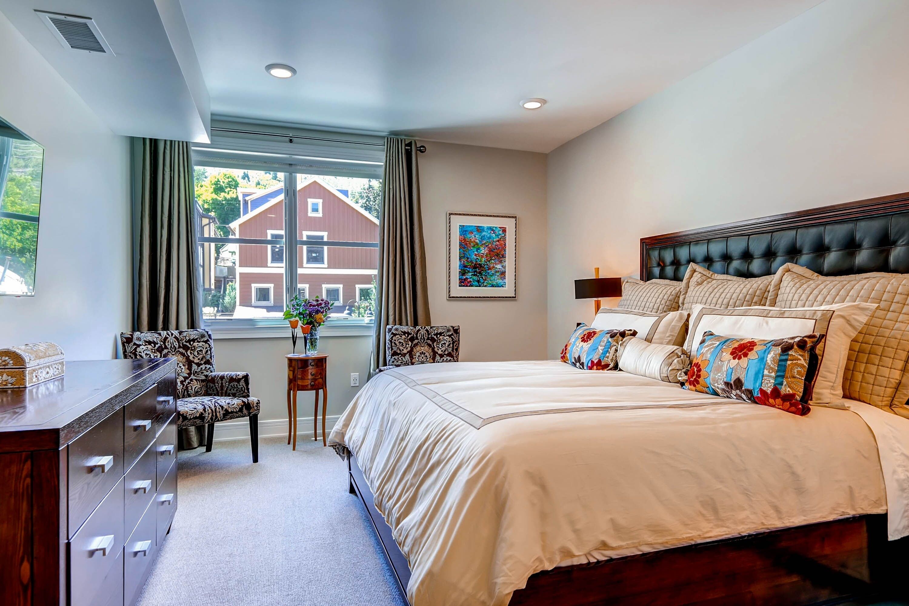 A bedroom with a large bed, a dresser, a chair, a window with curtains, and a painting on the wall.