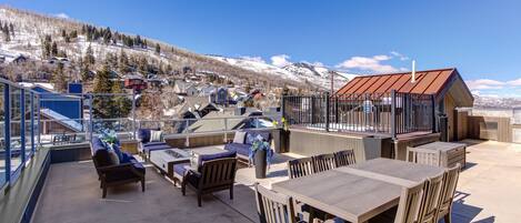 A rooftop patio with outdoor seating, a grill, and a view of a snowy mountain town.