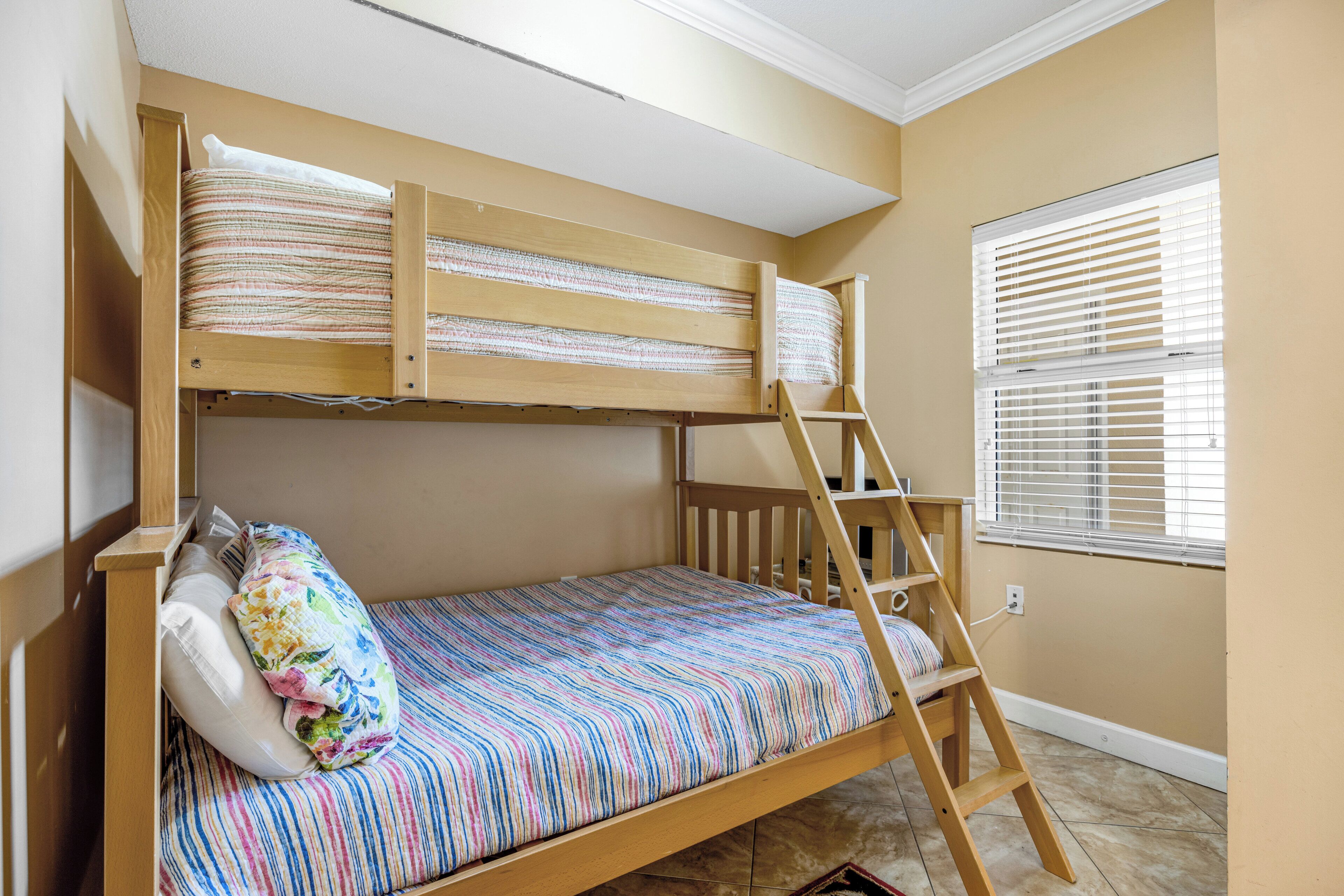 A bunk bed with a wooden ladder, a window with blinds, and a patterned bedspread.