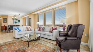 Amplia sala de estar con un sofá blanco, un sillón de cuero marrón y una mesa de centro de vidrio. Cuenta con ventanas grandes con persianas, un candelabro y una alfombra con estampado floral.