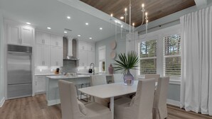 A modern kitchen with white cabinetry, a stainless steel refrigerator, and a dining area with a white table and chairs. - Designer Finishes, Arcade Room, Sleeps 14 & Next to Alys (Panama City Beach)