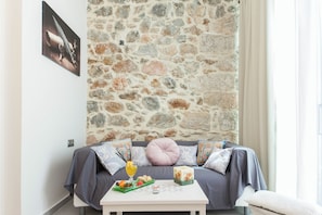 A cozy room with a stone wall, a bed with patterned pillows, a small table with snacks and a candle, and a framed picture on the wall. - Pireas Jewel New Design Apt w Backyard (Piraeus)