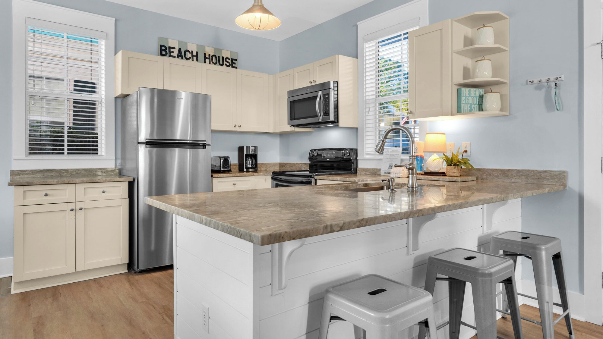 A modern kitchen with a stainless steel refrigerator, a microwave, and a stove. There are two bar stools and a countertop with a sink.