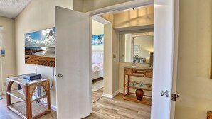 A room with a wooden floor, a glass-top table with books, a painting of a beach scene, a bathroom with a mirror, and a bed with a floral bedspread.