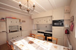 A compact kitchen with white cabinetry, a dining table with a patterned tablecloth, a chandelier, and a red kettle on the wall.