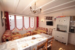 A cozy dining area with a long table, chairs, a chandelier, and a television on the wall.