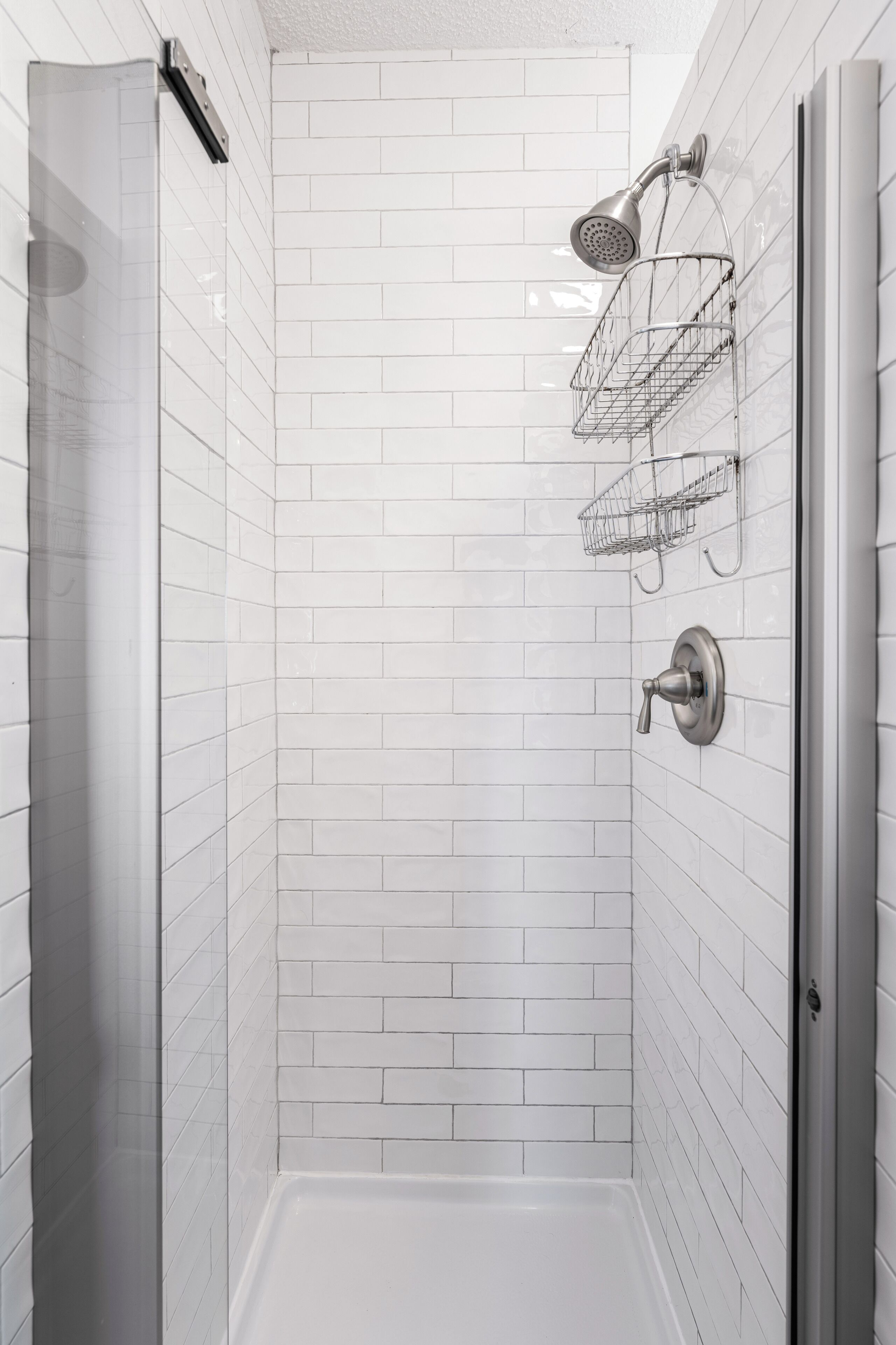 Une douche moderne avec des murs recouverts de carreaux blancs, une porte en verre et une pomme de douche fixée au mur.